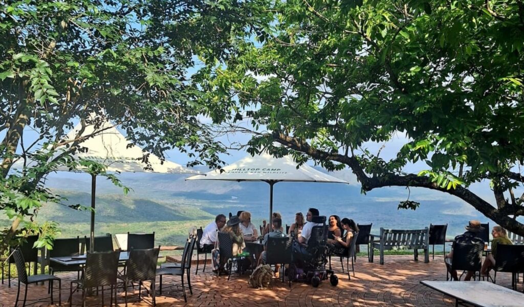 A wide shot of an outdoor patio at Hilltop Camp, featuring people gathered around tables under large white umbrellas. The patio overlooks a vast, verdant valley, with a person in a power wheelchair prominently seated at one of the tables, illustrating the site's accessibility.