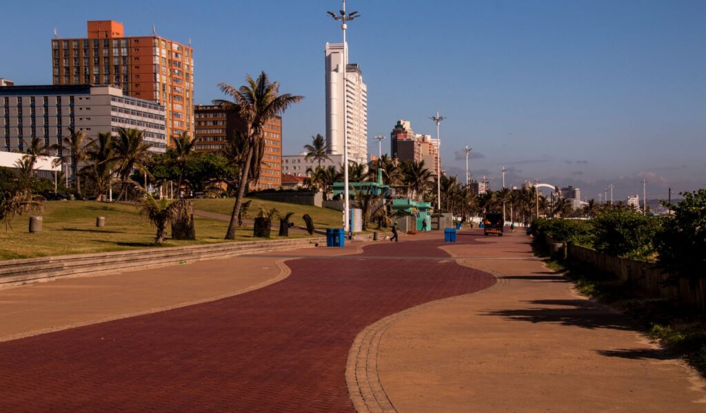 A sunny view of the Golden Mile promenade in Durban, South Africa, showing a wide red-brick walkway lined with palm trees, modern streetlights, and high-rise beachfront hotels against a clear blue sky.