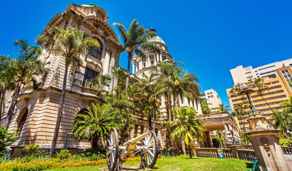 A historic sandstone building with domes and classical architectural details, surrounded by tall palm trees and lush gardens under a bright blue sky. A decorative old cannon and ornate lamp posts are visible in the foreground, suggesting a colonial-era or civic landmark.